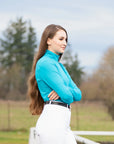 Woman in turquoise top and white pants standing outdoors with trees and a fence in the background