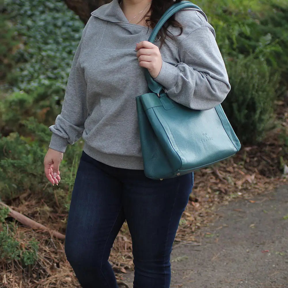 Person wearing a gray hoodie and blue jeans holding a teal handbag outdoors.