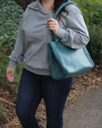 Person wearing a gray hoodie and blue jeans holding a teal handbag outdoors.