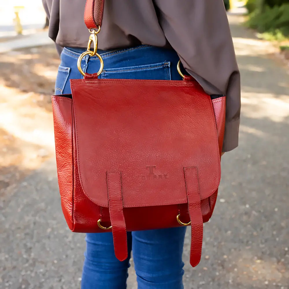 Red leather bag with gold hardware worn by a person on a blurred outdoor background