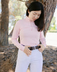 Woman wearing a pink long-sleeve top and white pants standing in a natural setting with trees.