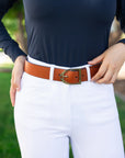 Person wearing a brown belt with a gold buckle on white pants, against a blurred green background.