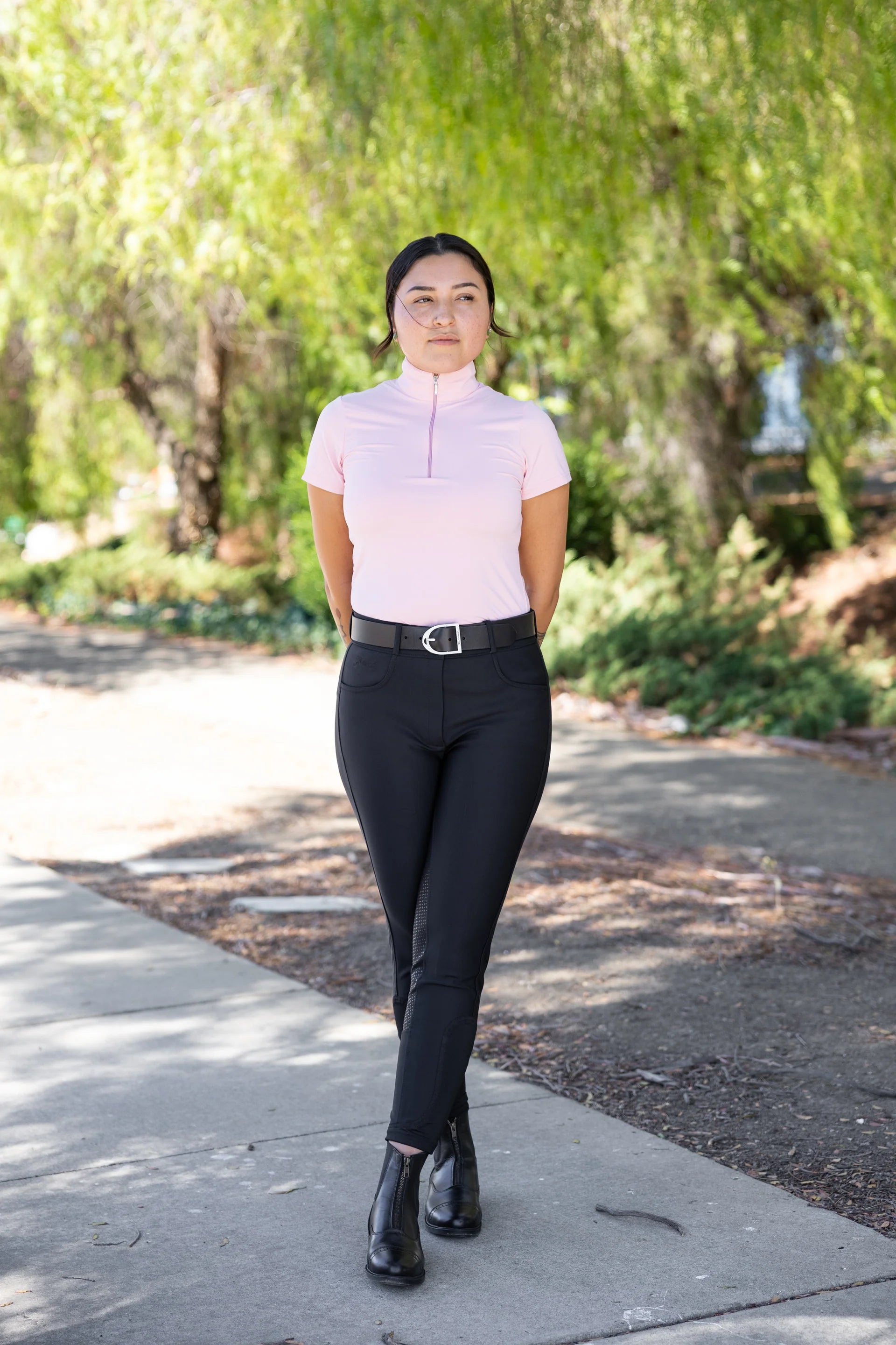 Woman in a light pink top and black pants standing on a sidewalk with trees in the background