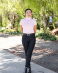 Woman in a light pink top and black pants standing on a sidewalk with trees in the background