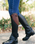 Person wearing black riding boots with brown accents and blue jeans outdoors.