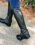 Black riding boots with gaiters worn by a person on a paved surface.