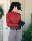 Person wearing a red long-sleeve top, black cap, and gray riding pants outdoors.