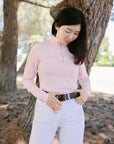 Woman wearing a pink long-sleeve top and white pants standing outdoors near trees.