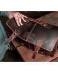Brown leather satchel bag held by a person on a wooden surface