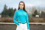 Woman wearing a turquoise equestrian top and white pants standing in an outdoor setting.