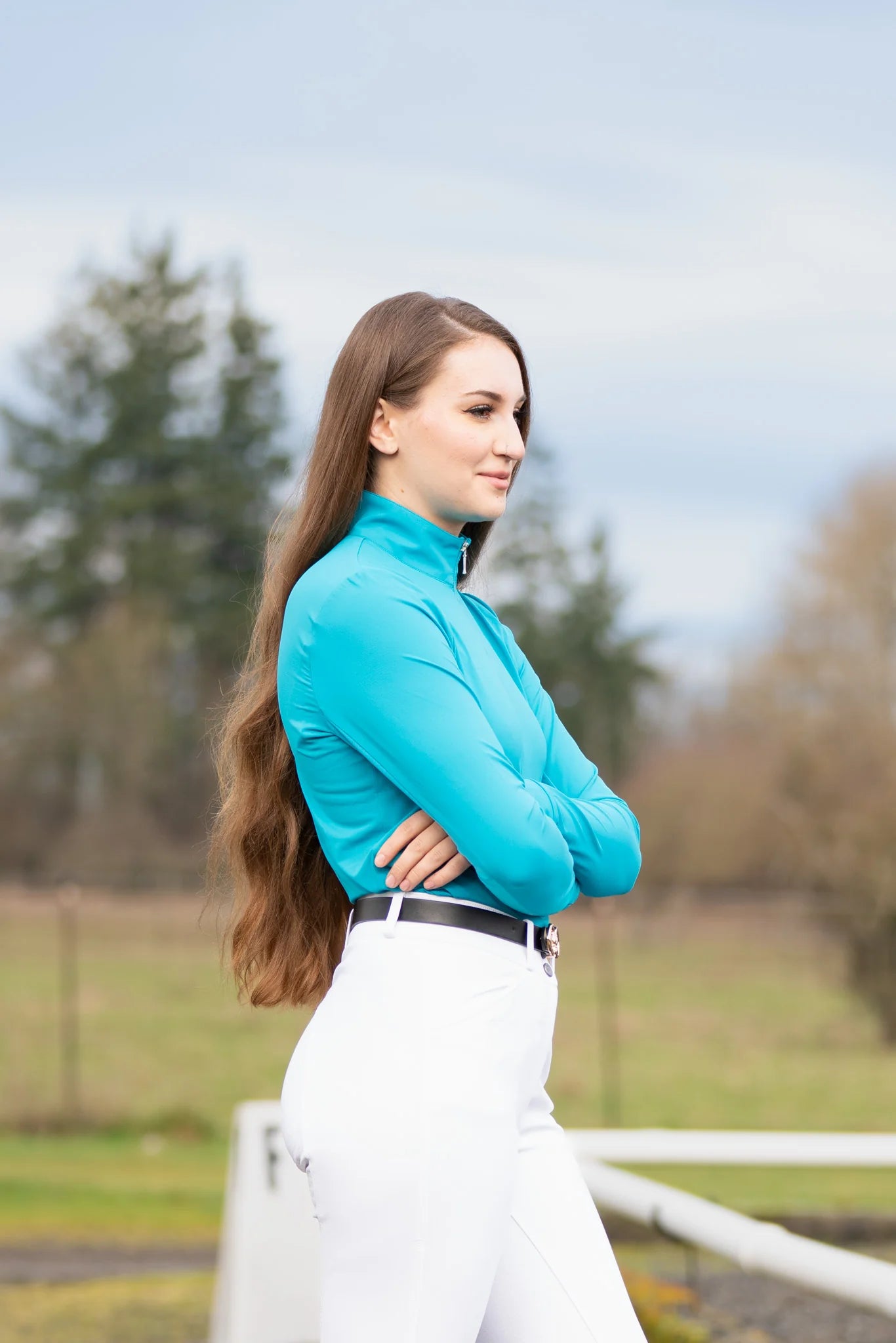 Woman in turquoise top and white pants standing outdoors with trees and a fence in the background