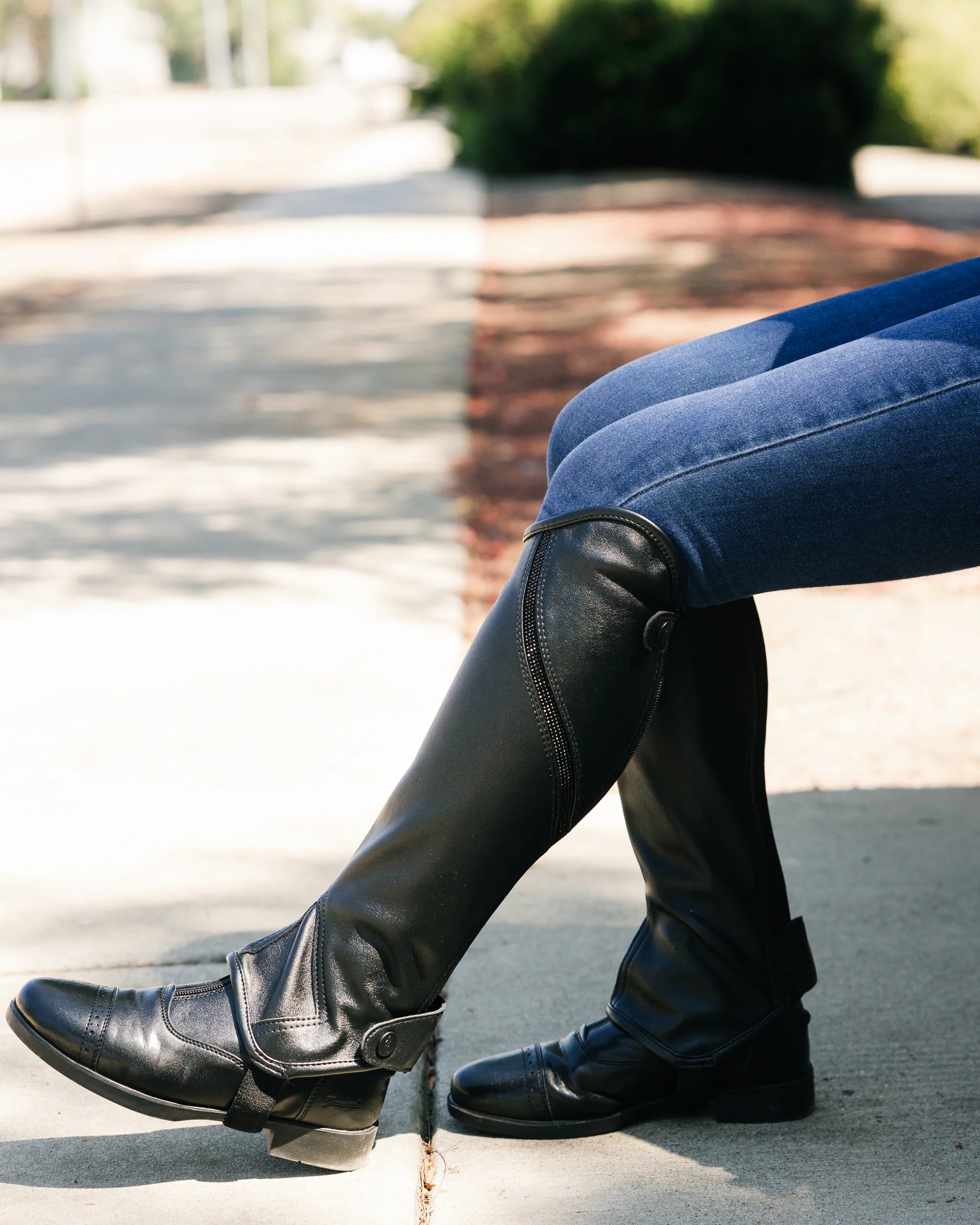 Person wearing black leather boots and blue jeans sitting on a curb.