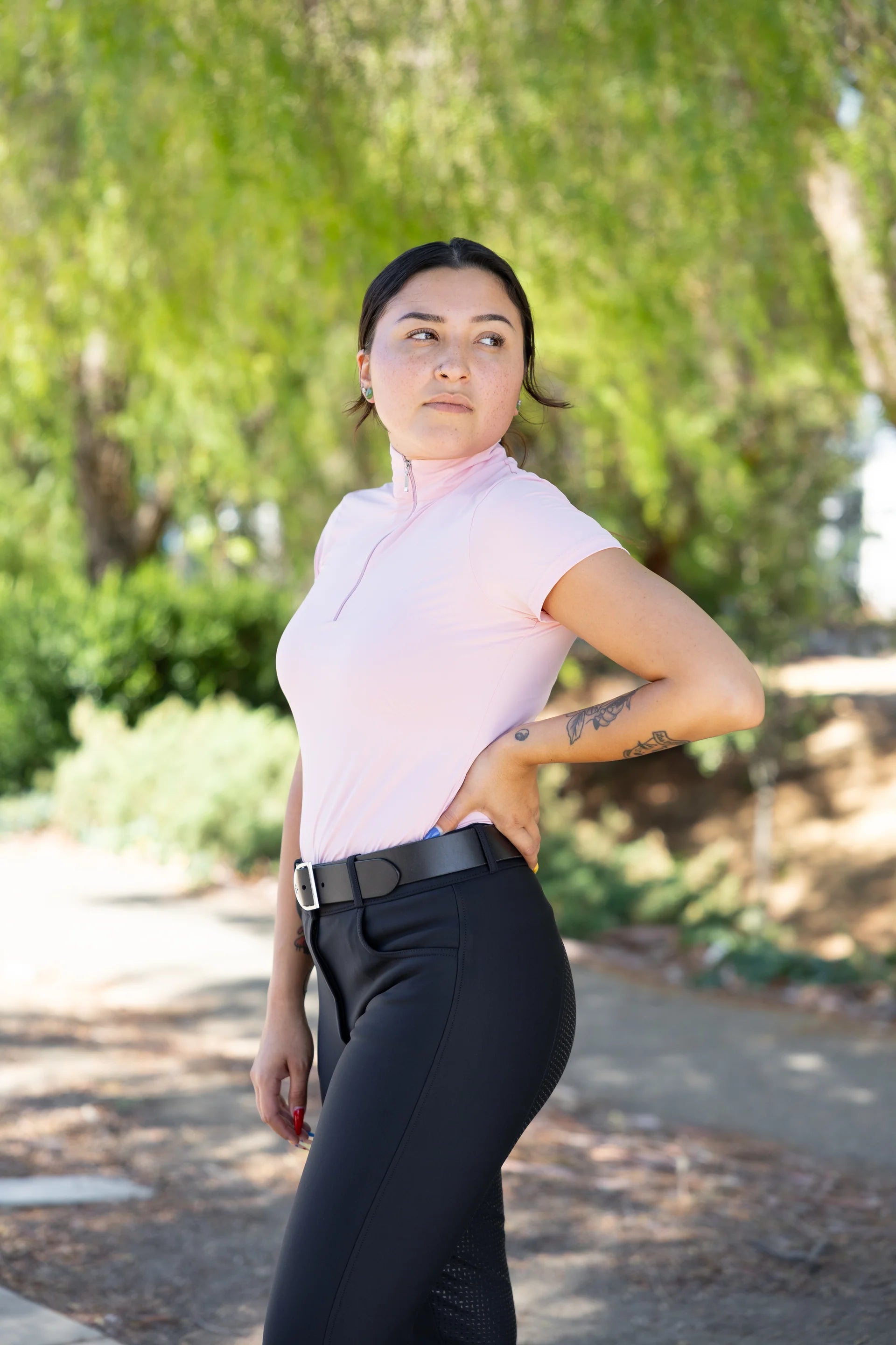 Person wearing a light pink shirt and black pants standing outdoors with greenery in the background